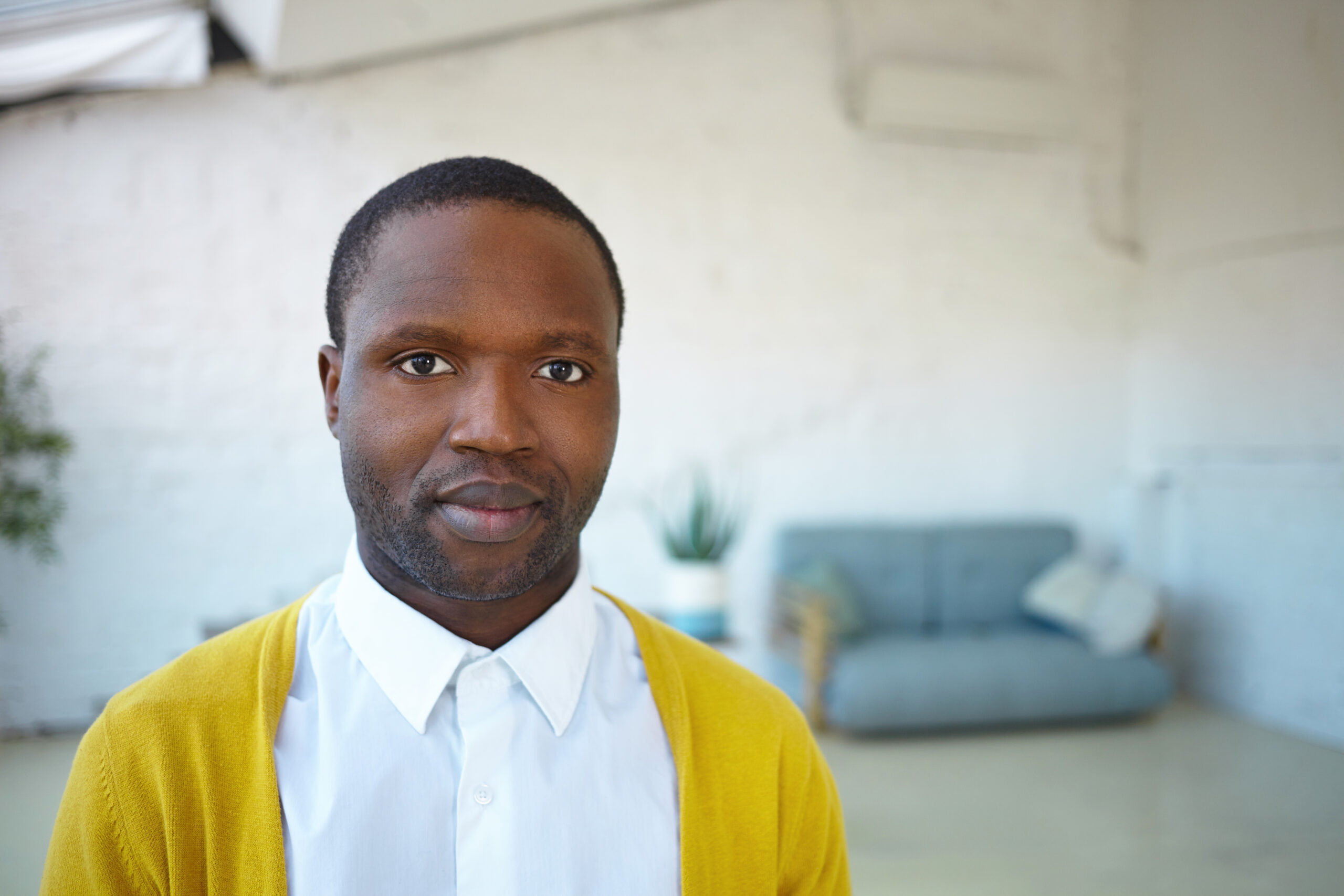Home close up shot of attractive young unshaven african american male wearing trendy yellow cardigan over white shirt, looking and smiling at camera, standing in modern living room with sofa in background